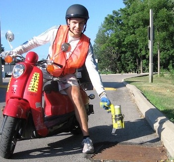 A picture of a person on a scooter treating a roadside catch basin to reduce the emergence of mosquito larvae.